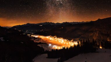 A breathtaking night sky filled with stars over a snow-covered valley with soft glowing lights from a nearby town.