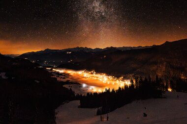 Ein atemberaubender Nachthimmel voller Sterne über einem schneebedeckten Tal mit sanften Lichtern einer nahegelegenen Stadt.