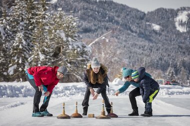 Drei Personen spielen Curling auf einem Freiluft-Eisbahn, umgeben von schneebedeckten Bäumen und Bergen.