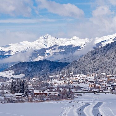 Eine Winterlandschaft mit schneebedeckten Bergen und einem Dorf unter einem blauen Himmel mit fluffigen Wolken.