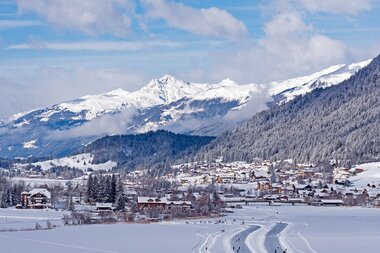 Eine Winterlandschaft mit schneebedeckten Bergen und einem Dorf unter einem blauen Himmel mit fluffigen Wolken.