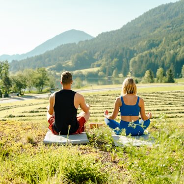 Ein Mann und eine Frau meditieren auf Matten in einer ruhigen Landschaft mit Bergen und Grün.