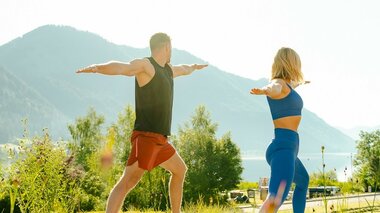 Ein Mann und eine Frau üben Yoga im Freien vor einer Berglandschaft.