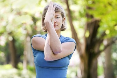 Eine Frau in einem blauen Shirt macht eine Dehnung, mit einem Arm, der über ihr Gesicht gekreuzt ist, in einem grünen Park.
