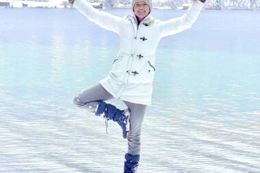 Eine Person, die einbeinig an einem schneebedeckten See im Winter steht, trägt einen weißen Mantel und eine gestrickte Mütze.