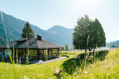 A picturesque house surrounded by grass, trees, and mountains in bright sunshine.