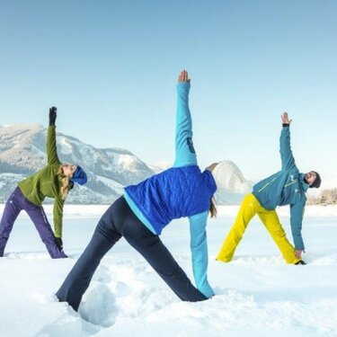 Three people in winter clothing performing yoga poses in the snow with mountains in the background.
